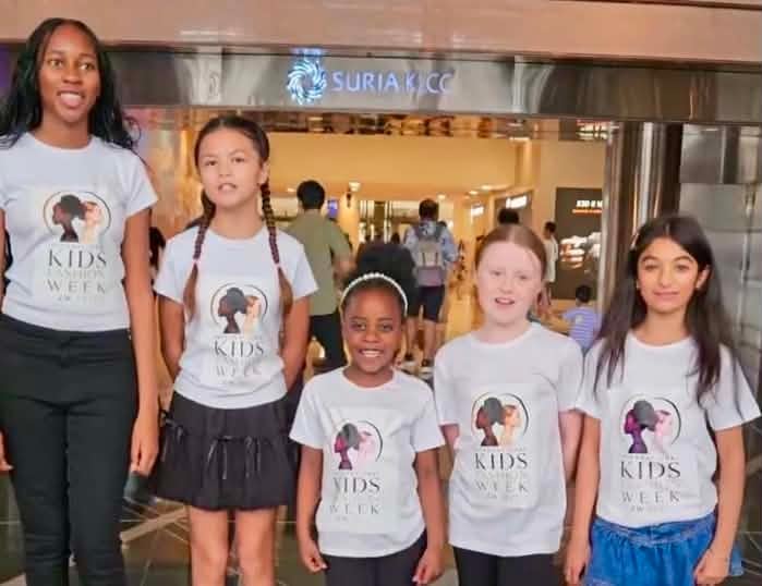 Five diverse children wearing matching Kids Week t-shirts pose together in a shopping mall corridor
